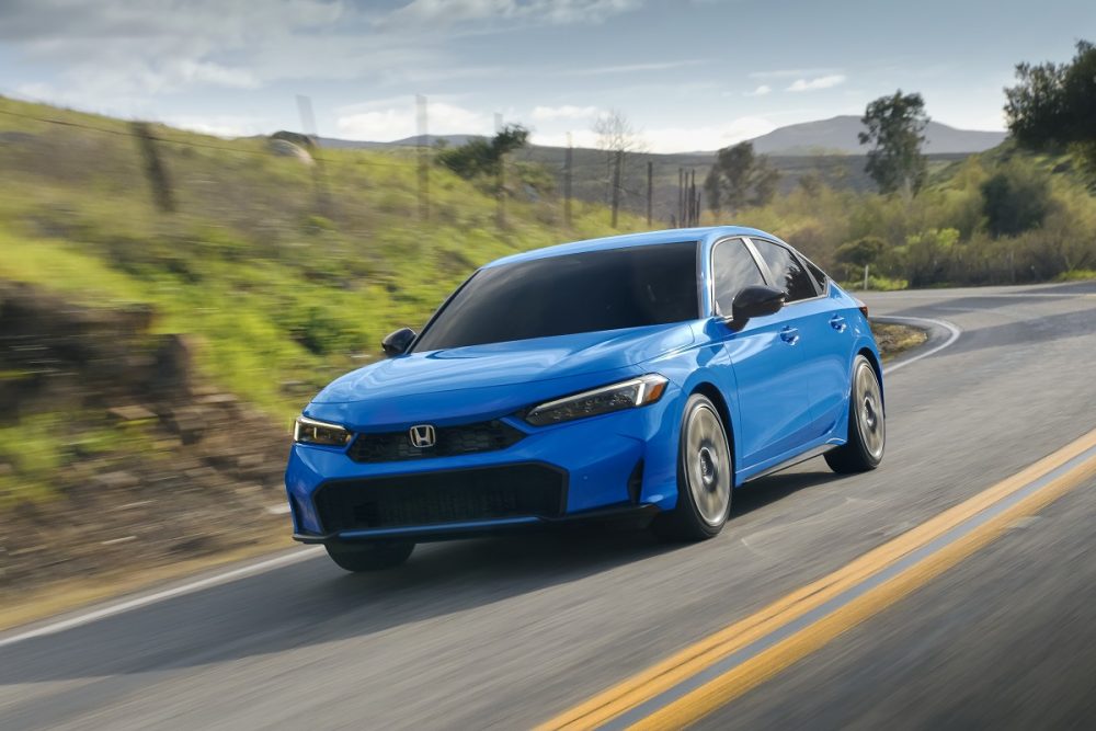 Front side view of a blue 2025 Honda Civic Hatchback Sport Touring Hybrid driving down a road