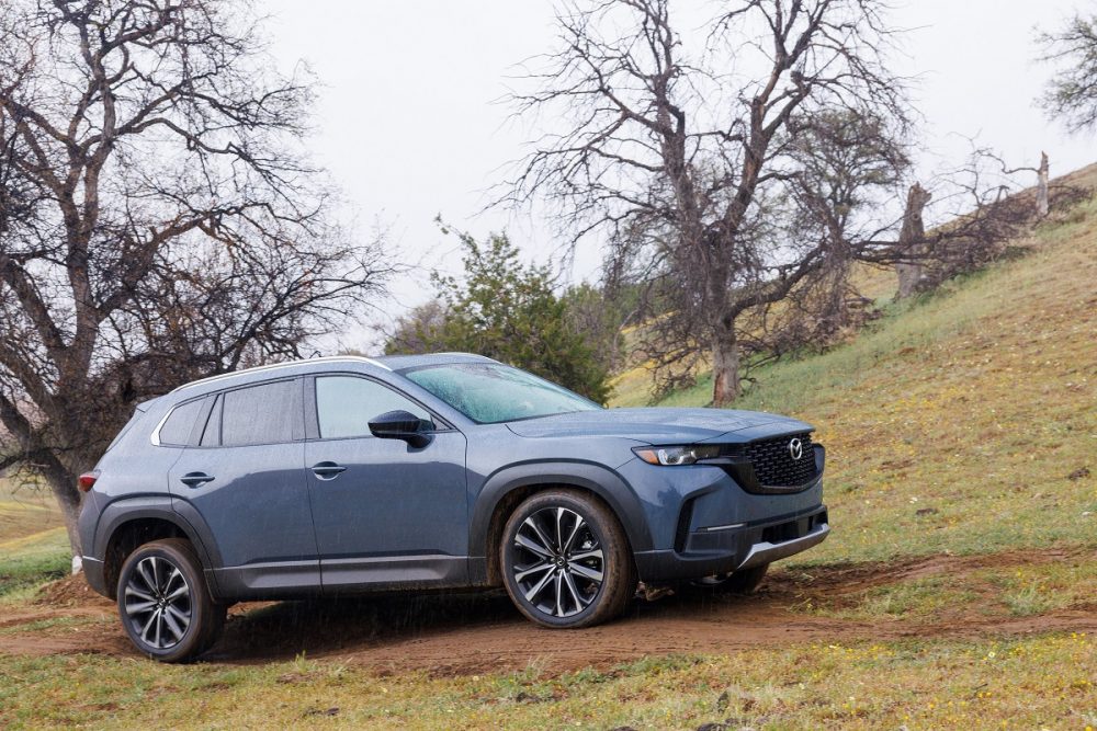 A side view of the 2025 Mazda CX-50 driving up a muddy, tree-studded hill in the rain