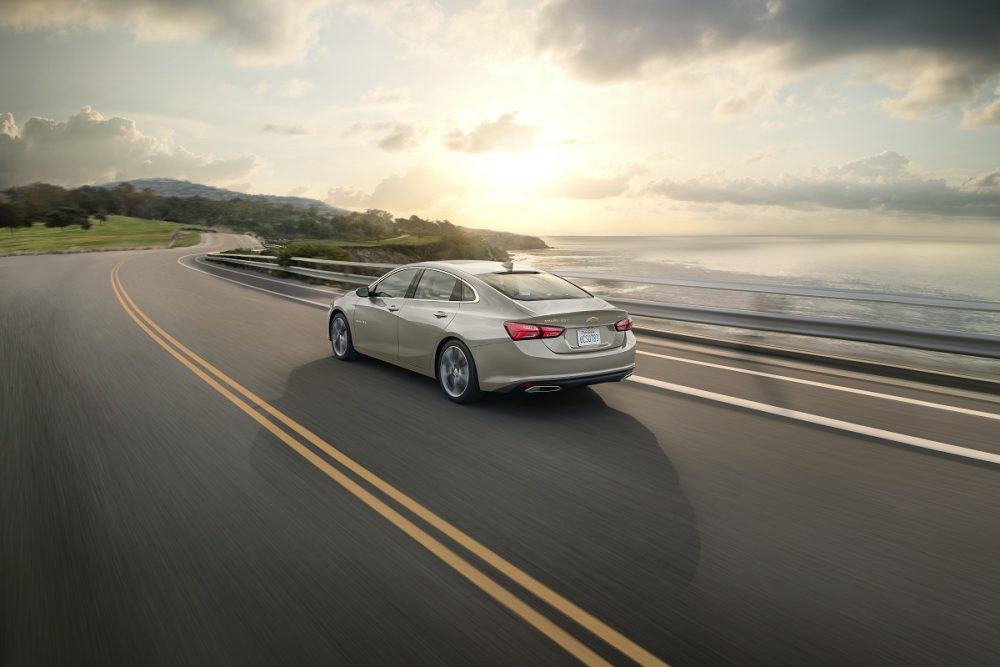 Rear 3/4 view of the 2024 Chevrolet Malibu driving near a body of water with the sun low in the sky